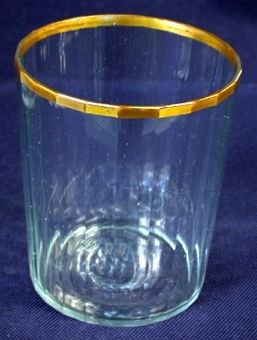 Cylinderformat dryckesglas av genomskinligt glas. Öppningens kant förgylld som en guldbård som löper runt öppningen. Utsidans yta har ett långrandigt mönster. I botten en långsmal figur i relief.