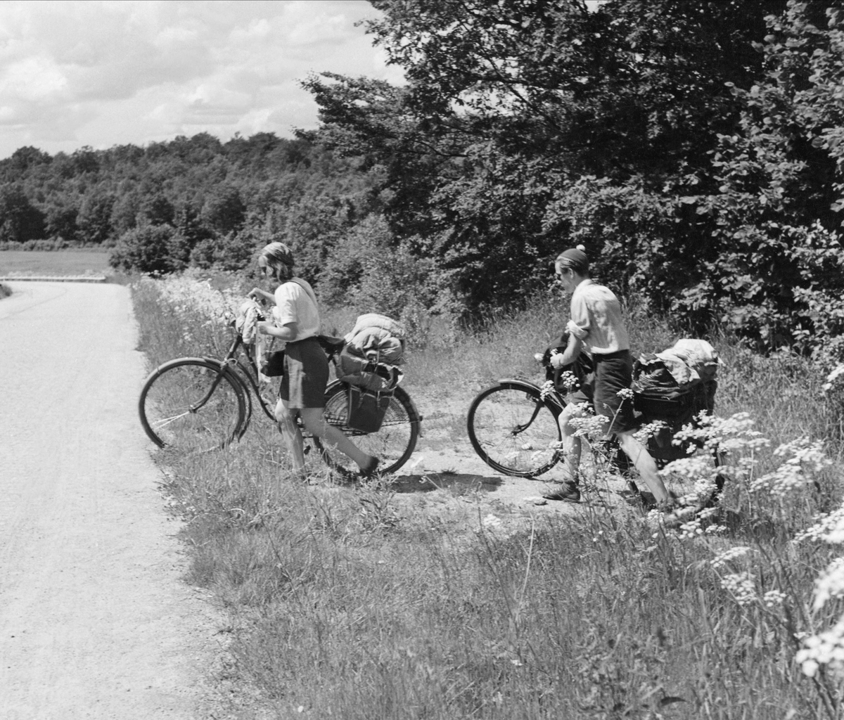 Två cyklister i skogslandskap, ung kvinna och ung man, leder cyklar med stor packning från vägkanten och ut på en grusväg.