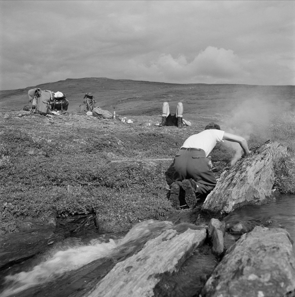 Dag Hammarskjöld på fjällvandring på Sulitelma. Två män har slagit läger, den ene ligger på rygg och vilar medan den andre gör upp eld vid ett klippblock.