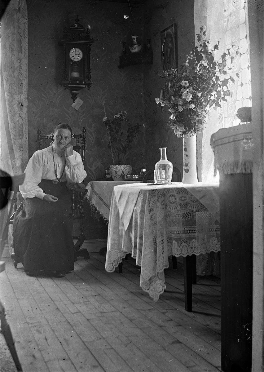 Småskollärarinnan Bertha Engström i Sällstorp sitter i en gungstol i lärarbostadens finrum. Över henne hänger ett väggur som visar på klockan tre, och på bordet står en karaff med dricksglas på bricka och en hög vas med stor bukett.