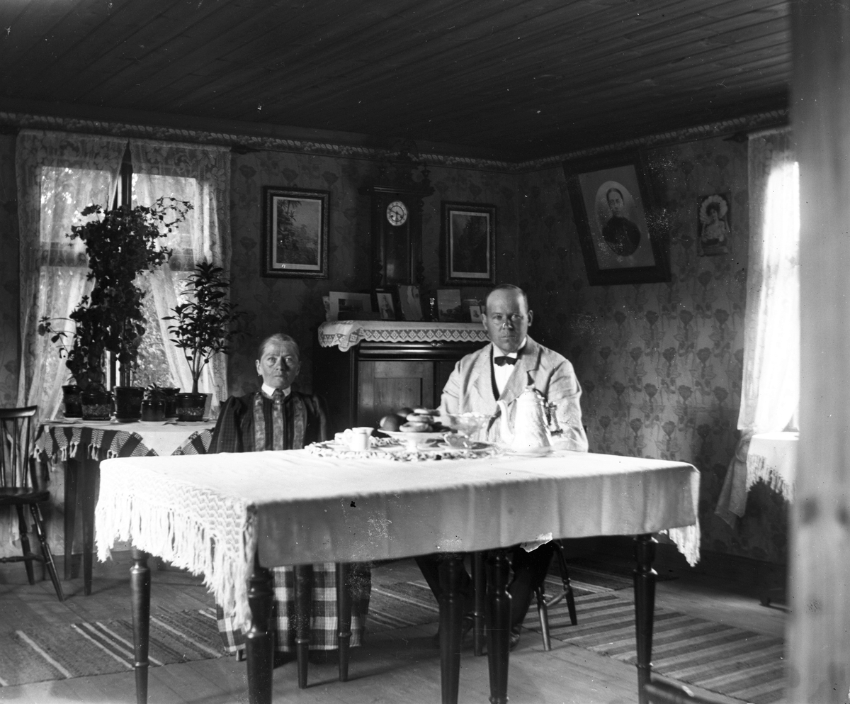 En man och en liten äldre kvinna i rutig kjol sitter vid ett stort bord i finrummet. På bordet står en bricka med "doppe" (bakverk), sockerskål, mjölkkanna och kaffekoppar. De blommiga jugendtapeterna har en bård upptill.