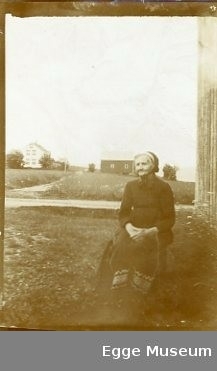 Kristianne Nilsdtr. Mortensen (1823-1918) sitter på en stol/krakk utenfor en bygning. 