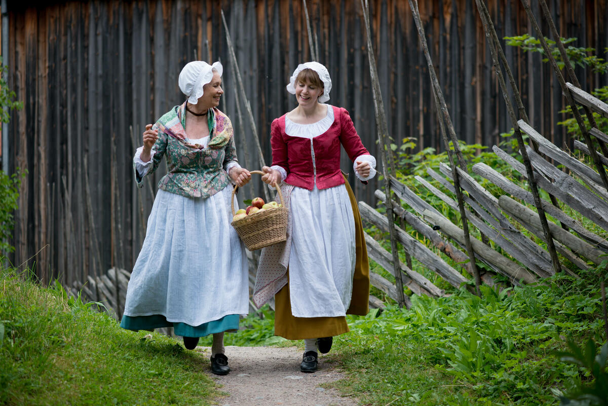 To kvinner i gammeldagse klær kommer gående på en sti med kurv i hånden.