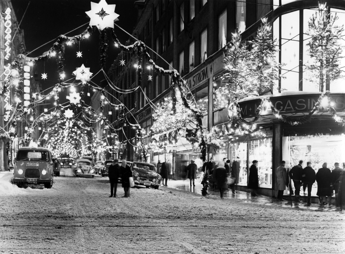 Foto av julehandel i Oslo.Gata utenfor Steen og Strøm er julepyntet. Biler står parkert i Nedre Slottsgate.