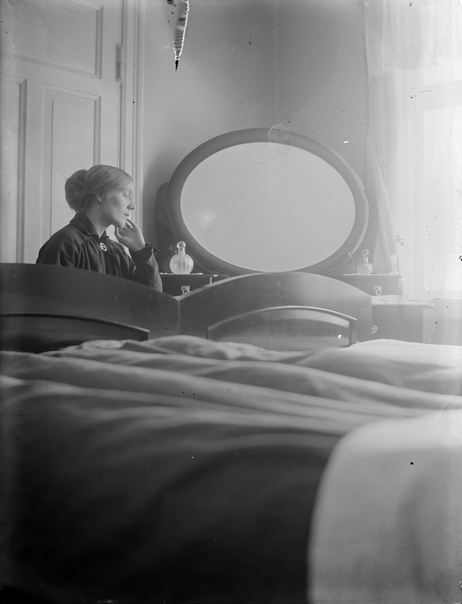 Portrett av en kvinne på et soverom. Bbildet er tatt fra en lav vinkel, ei seng dekker hele nederste delen av bildet. Kvinnen sitter ved et speil og er belyst av et vindu på høyre side.