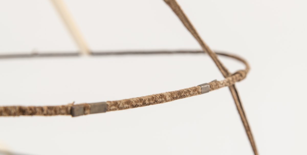 Krinoline med metallstativ knyttet sammen med brikkevevet bånd i bomull. Jernringene er beskyttet med bomullsbånd. Ser ut til å mangle 3 ringer. Krinolinen har et bånd festet i livet som festets sammen med beltespenne.
