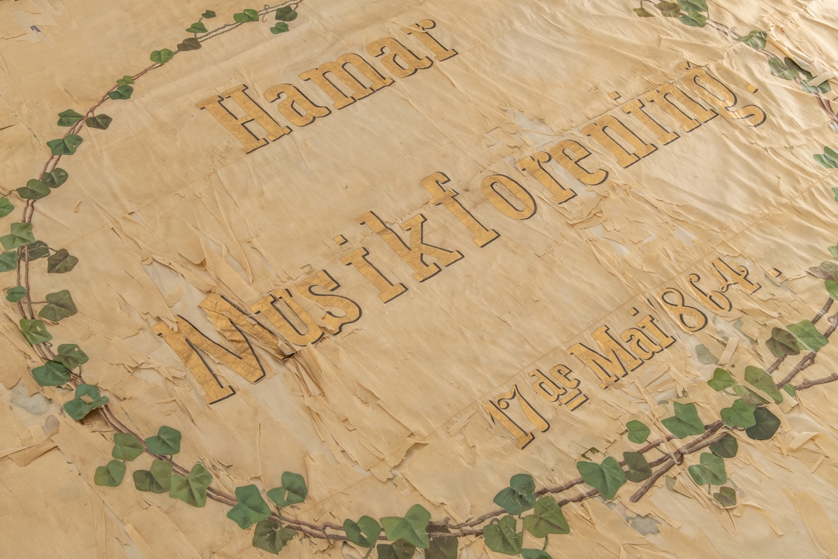 Fane for Hamar Musikkforening. Malt sirkel med blomsterranker rundt den påmalte teksten: "Hamar Musikforening. 17de Mai 1864" på framsiden og påmalt blomsterranke i sirkel rundt engel som blåser i valthorn på baksiden.