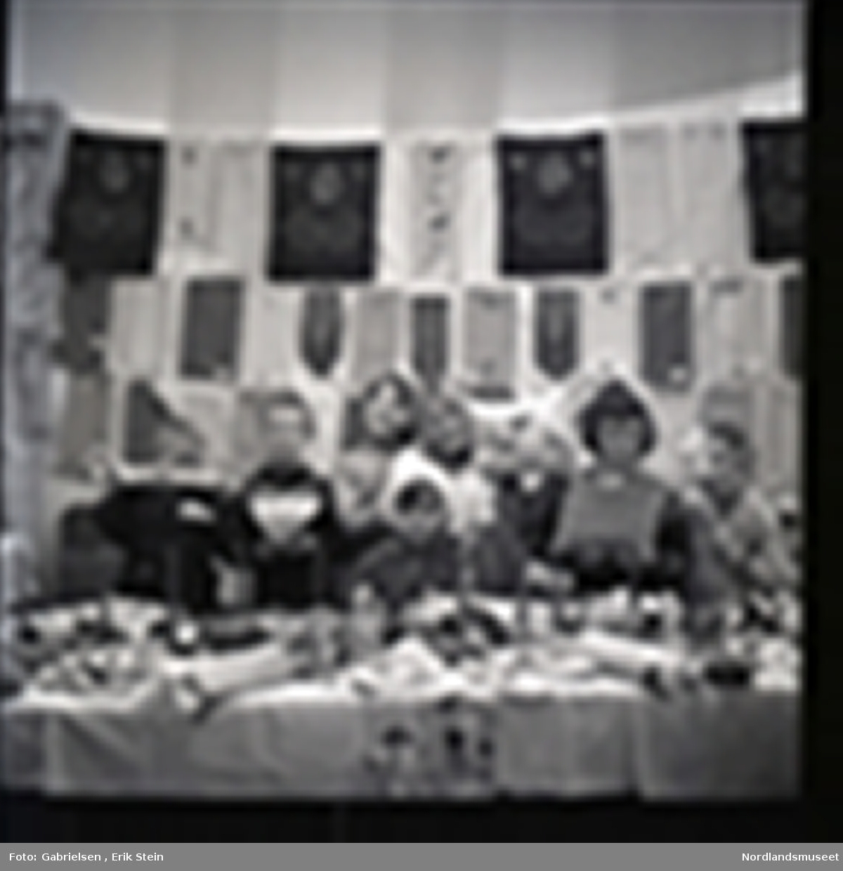 Fotografi av en gruppe med barn som selger puter med mønster og julelys og kurver og kjevler og juleduker og noen tøfler og andre ting på et langt bord i et rom med et vindu med noen gardiner et ukjent sted