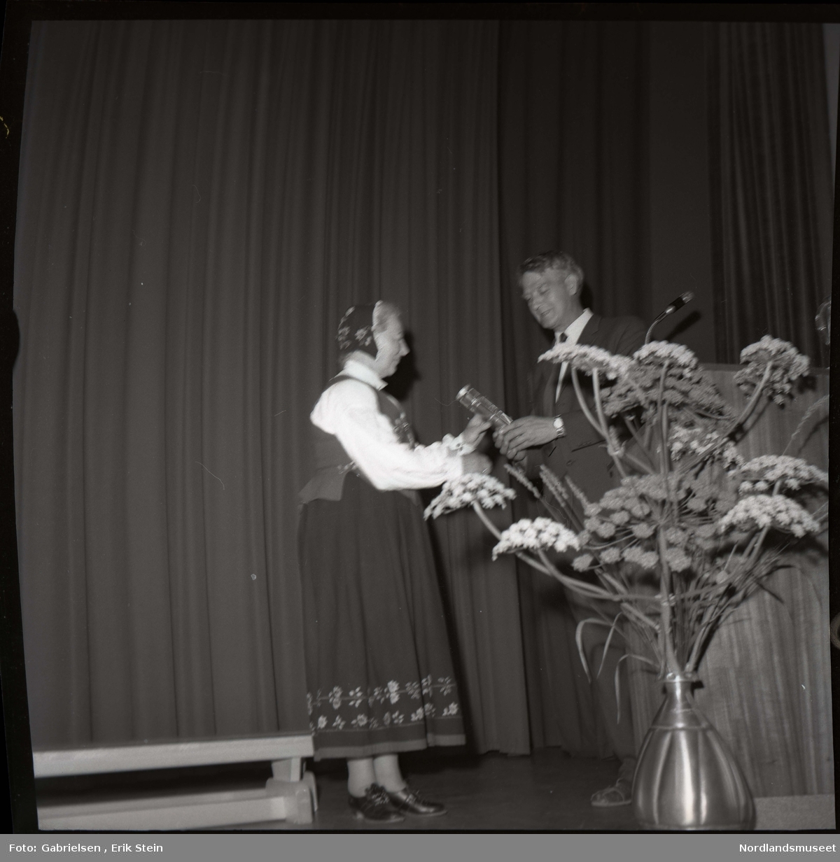Fotografi av en eldre kvinne kledd i en bunad og en bunadshette som står på en scene i en sal og får utdelt en pris ev en mann kledd i dress med slips som står vedsiden av en talestol med en mikrofon og en stor blomsterkrukke 
som det vokser mange blomster i og en benk som står på scenen og man ser en rekke av blomstermønster på
bunaden til kvinnen i skjørtet på bunaden og bunadshetten