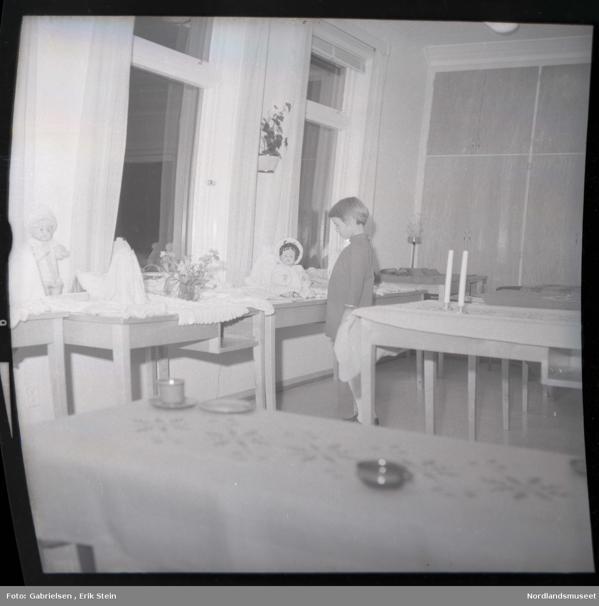 Fotografi av et barn som står inne i et rom og ser på en dukke
som ligger på en bord med en hvit duk 
som står mellom fire bord som står vedsiden av vinduer med gardiner som det ligger hvite duker på og en dukke som står lent på en vegg mellom to vinduer og man ser to andre bord
som står i rommet med duker på og to stearinlys som står
på bordet som står bak barnet i rommet og man ser
en potteplante som henger på veggen mellom to vinduer