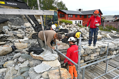 Bildet viser arbeidere som jobber med å bygge opp en større mur. I bakgrunnen ser man et rødt hus og grå himmel.. Foto/Photo
