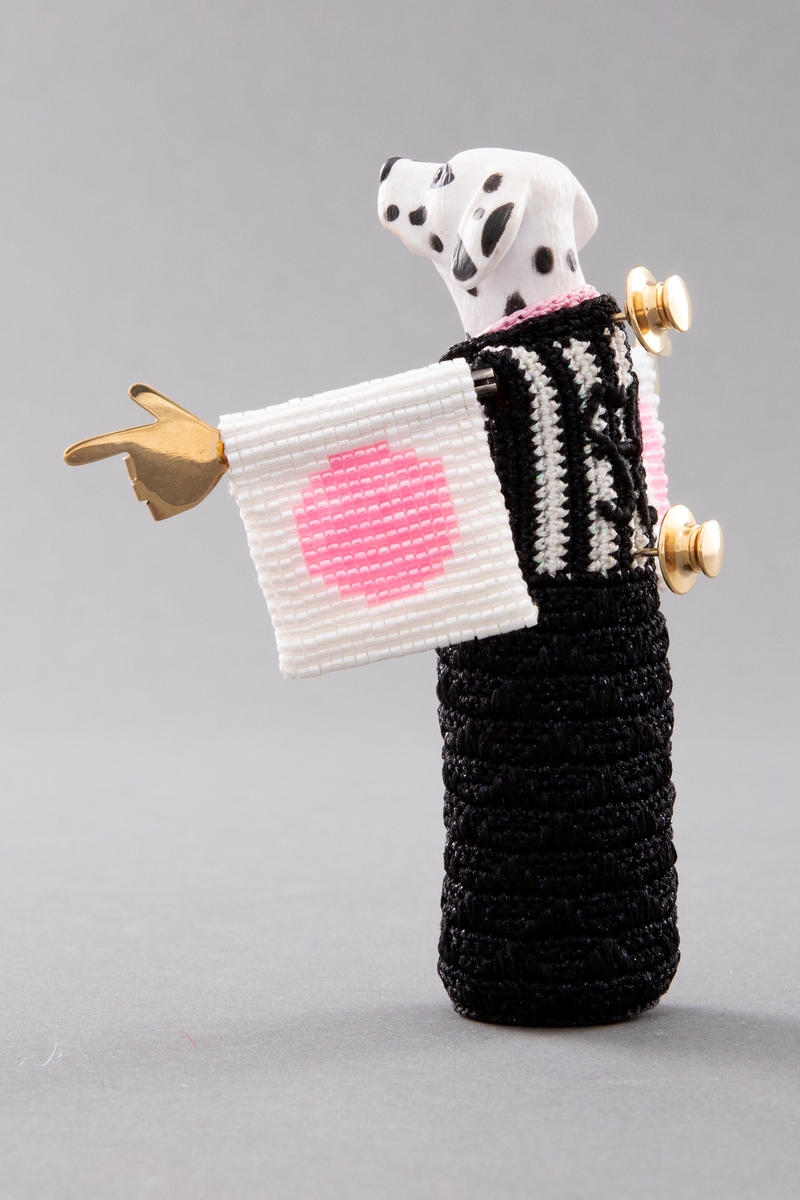 Brosje laget av en bearbeidet leketøy-dalmatinerhund i plast. Figuren er ikledd en heklet svart kimono med hvite vertikale striper på overdelen og har rosa halspynt. Kimonoermene er av perlebroderi i hvitt med en rosa sirkel på for- og baksiden. Figuren har svarte øyne av kubisk zirkonia, bevegelige armer og gullhender med pekende fingre. Brodert signatur i svart tråd og to festepins i metall på baksiden.