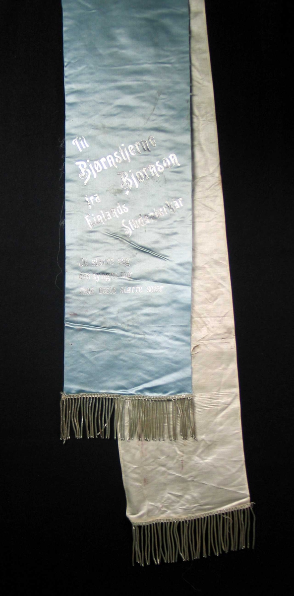 Begravelsessløyfe i hvit og blå silke med frynser og tekst i sølv.
