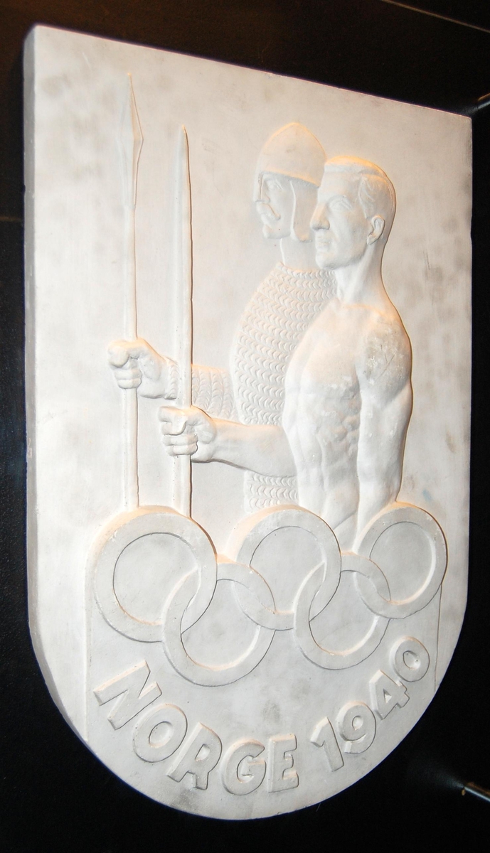 Skjoldformet plakett av gips med motiv av to menn, en i rustning og hjelm og en i bar overkropp. De olympiske ringene inngår i motivet.