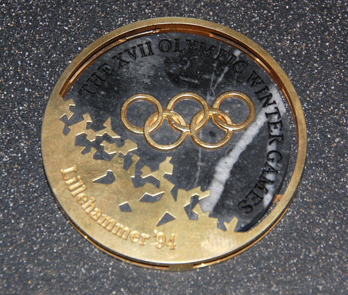 Medalje av metall og stein. På den ene siden er det motiv av de olympiske ringene over krystallmønster. På den andre siden er det piktogram av en skøyteløper og emblemet for de olympiske vinterleker på Lillehammer i 1994. Medaljen er plassert på et stativ av granitt.