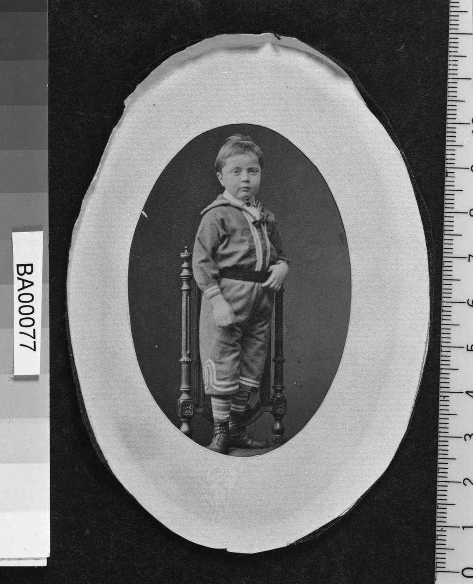 Fotografi av gutt stående på stolsete, kledd i matros drakt med bredt mørkt belte om livet, stripete strømper og støvler. Han ser direkte på betrakteren.