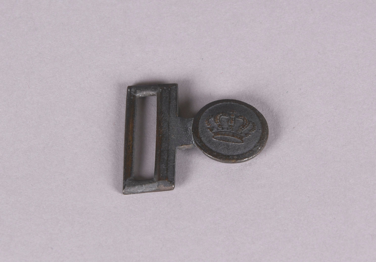 Beltespenne av kobberlegering. Rektangulær spenne med avlang åpning, rund knapp med emblem som muligens tilhører det norske forsvaret.