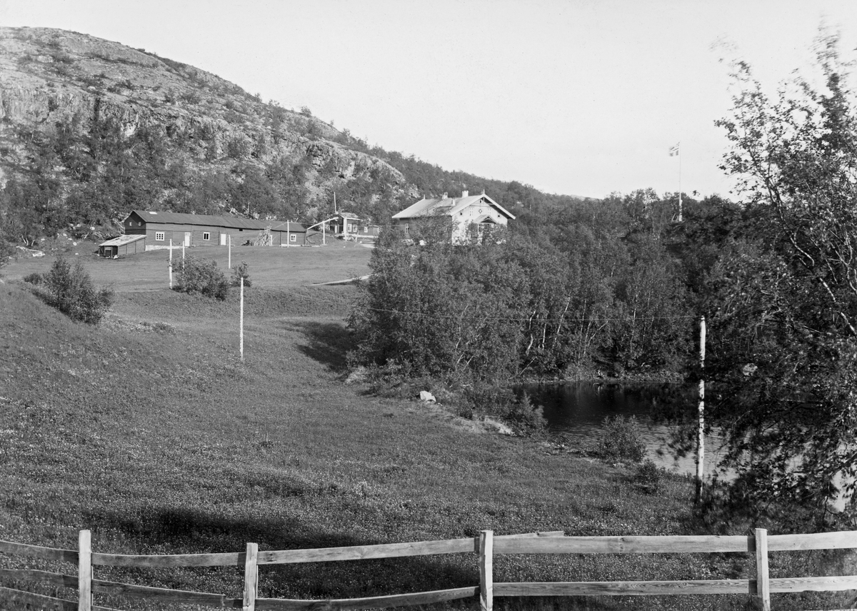 Garden Elvenes i Sør-Varanger.  Eiendommen tilhørte skogforvalter Arthur Klerck (1866-1943) i Øst-Finnmark Skogforvaltning, men ble i 1920 overtatt av det statlige skogvesenet som embetsbolig for etterfølgerne i embetet.  Garden er fotografert nedenfra med et bordgjerde mot et engareal i forgrunnen.  Til høyre i forgrunnen skimtes dessuten en vannbredd omgitt av lauvskog.  I gardstunet ses (fra venstre) en lang, rødmalt driftsbygning, en liten sidebygning og et halvannenetasjes våningshus med sveitserstilpreg.  I bakgrunnen et berg med spredte bjørkekratt. 