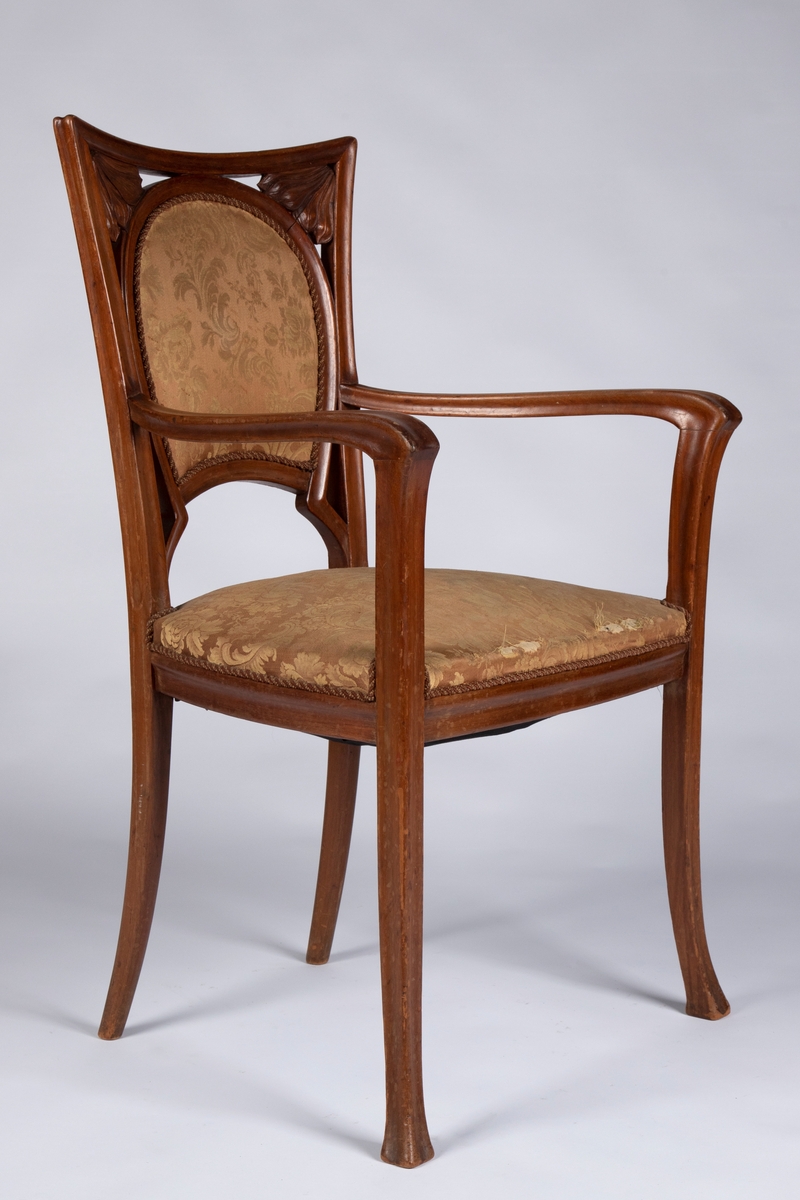 Armstol i tre, som hviler på fire slanke ben. Forbenene er utformet som kurvede plantestengler, og glir over i stolens armlenstøtter og buede armlener. De kurvede bakenene glir over i ryggstolpene, som innrammer et skjoldformet, gjennombrutt ryggbrett. Konkavt toppstykke, hvor overgangene til ryggstolpene er markert med blomsterornamenter i høyt relieff.  Setet er trukket i gul silke.
