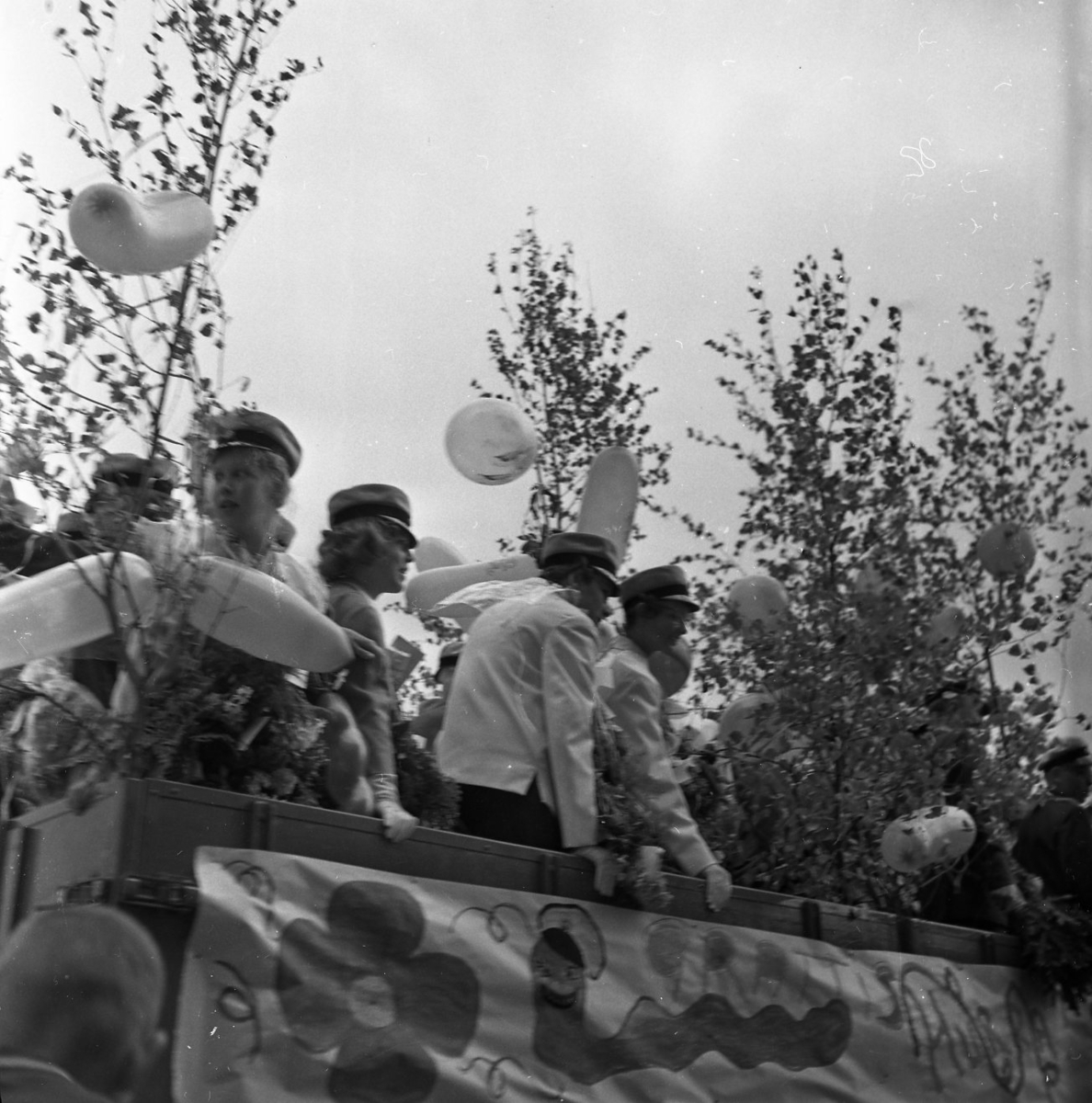 Realexamen 1961. Bilden föreställer ett lastbilsflak, på flaket står flera studenter. De bär studentmössa och blombuketter runt sina halsar. Flaket är klätt med Björkris och ballonger.