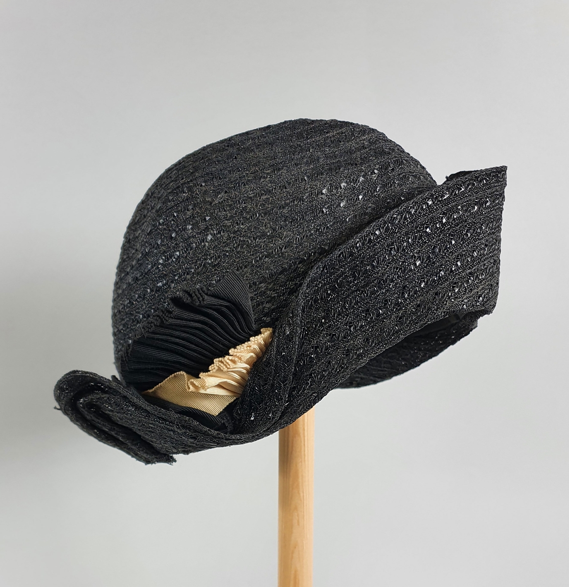 Koksgrå damehatt av strå, med høy pull og brem som er brettet opp foran og langs sidene. Svart hattebånd rundt pullen som ender i en liten vifte på høyre side.