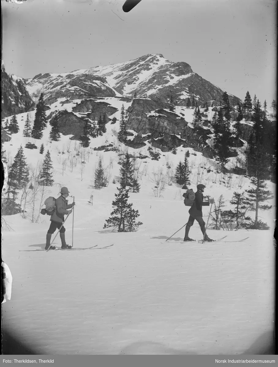 To menn med sekk på ryggen går på ski i Blefjell. Fjell i bakgrunnen.
