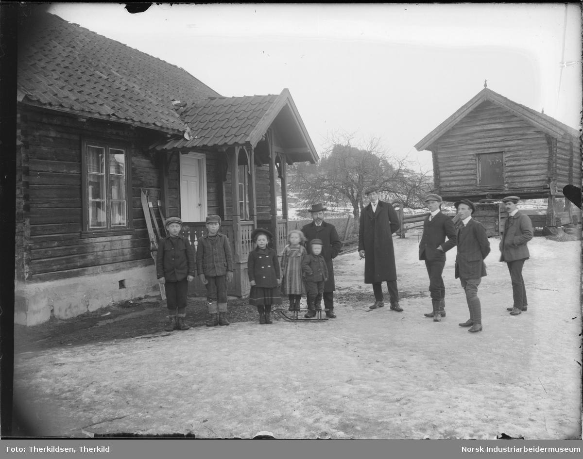 Ti mennesker stående på gårdstun på Sagafoss husmannsplass. Fem barn og fem menn står ved inngangen til bolighuset. Ski står opp mot veggen på huset. Stabbur bak mennene.