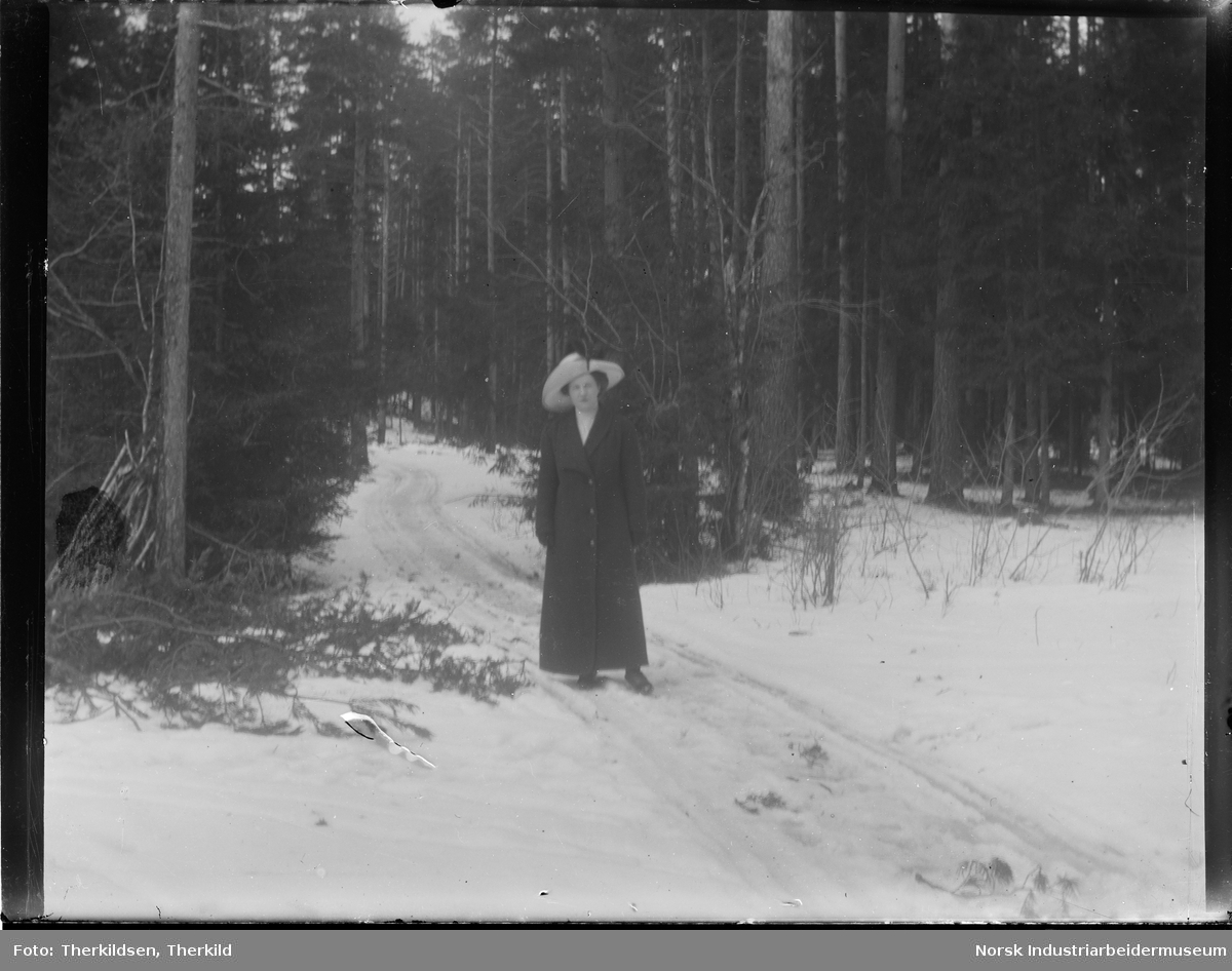 Emilie Knutsen stående på snødekket sti gjennom skogsterreng. Kledd i kåpe og hatt.