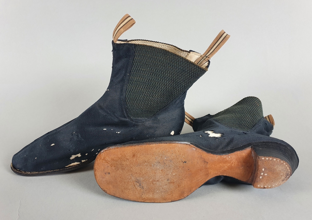 Ankelhøye, smale, svarte sko av tekstil. Lav hæl. Innfelt grovt vevet tekstil på begge sider av ankelen. Stropp foran og bak. Lærsåle, den ene er løsniet fra sålen.