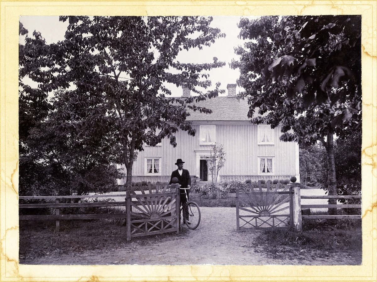 En man med cykel står vid grinden, framför en (trol. gulmålad) mangårdsbyggnad.
Fålaboda, Urshult.