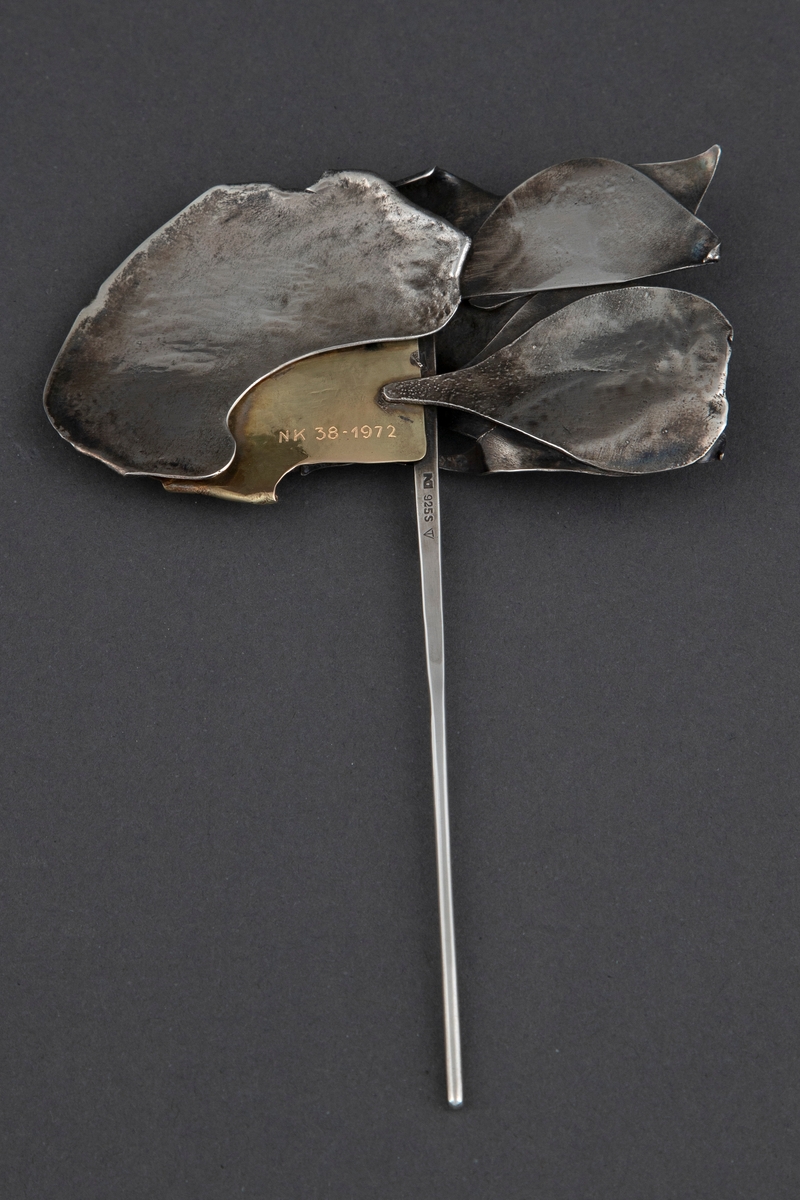 Brosje med stiliserte bladformer av smidd sølv, samt en gullplate, festet til en lang nål. Sølvet har en ru, matt overflate, mens gullet er blankt.