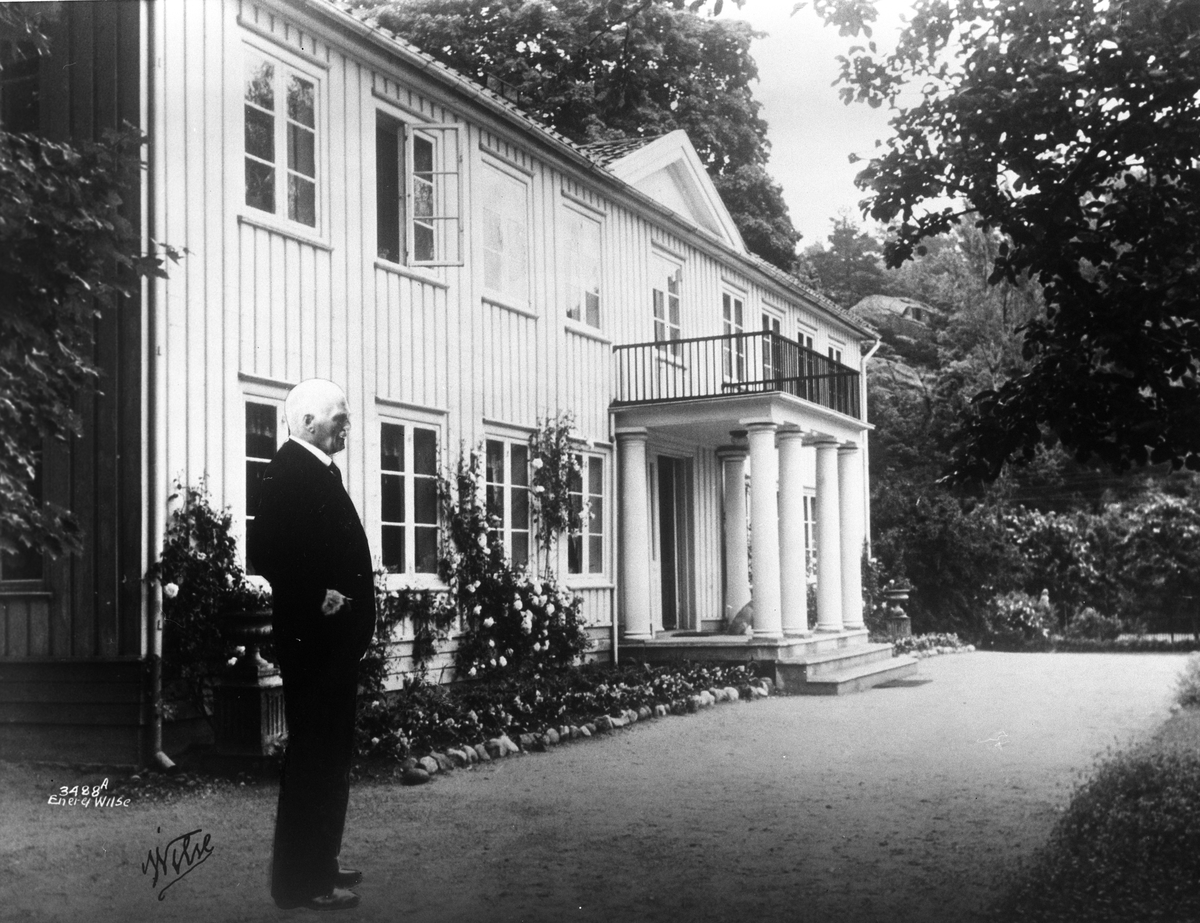 Knut Hamsun står utenfor husets inngangsparti med søyler og trapper. Et vakkert hus og parkanlegg.