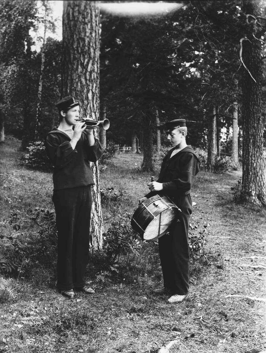 2 marinegaster fra Sjøkrigsskolen spiller på sine instrumenter, trompet og tambur ute i et hyggelig skogsterreng.