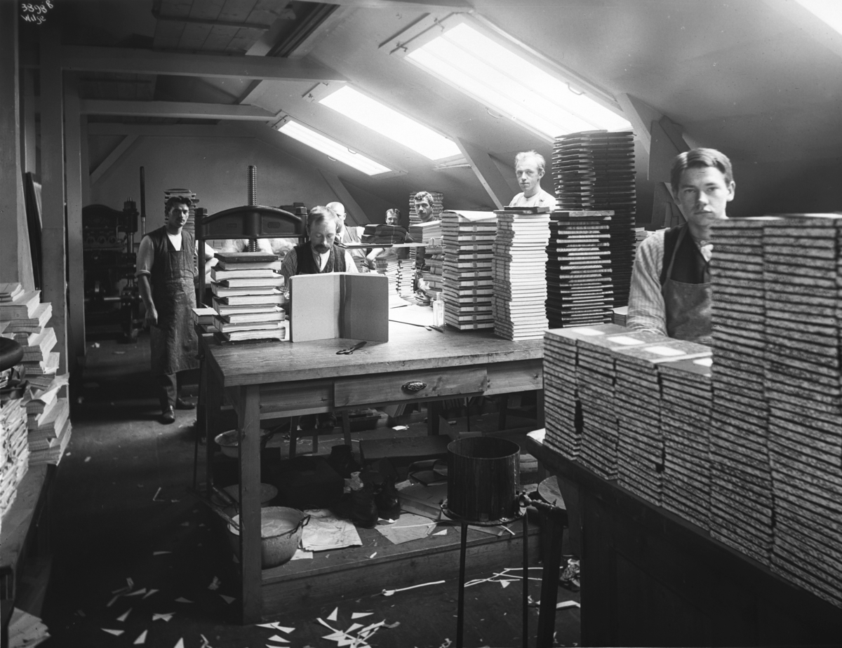 Bokbinderfaget, et gammelt håndverk. Et blikk inn i verkstedet med ferdige produkter og en gruppe menn i arbeid.