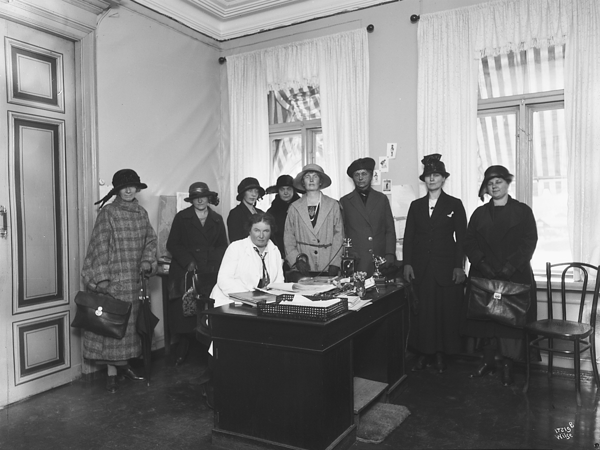 En dame sitter ved skrivebord, flere damer står rundt, Kvia-kommunernes pensionretning. Fotografert 1923.