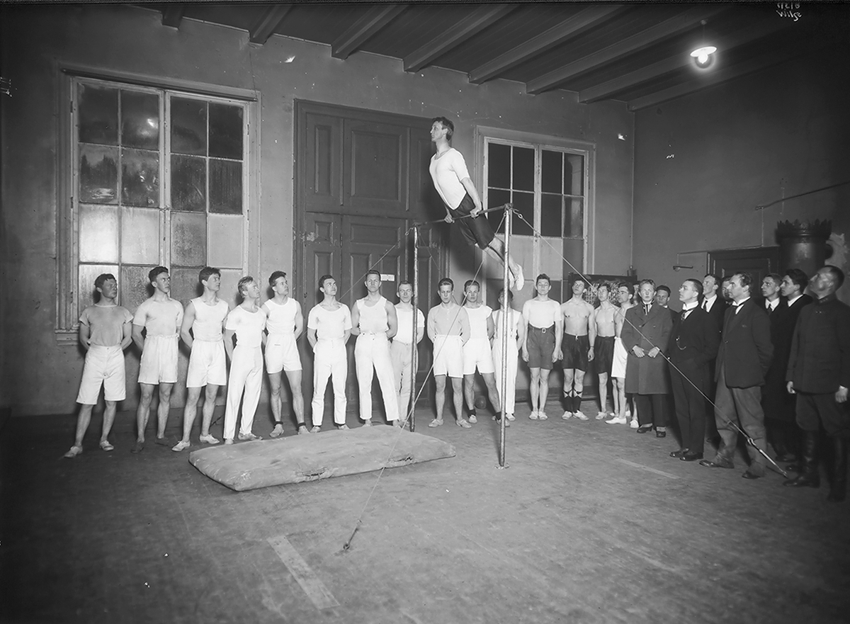 Turnere står sammen med menn i dress og ser på en mann i svingstang, Studentenes Gymnastikksal. Fotografert 1923.