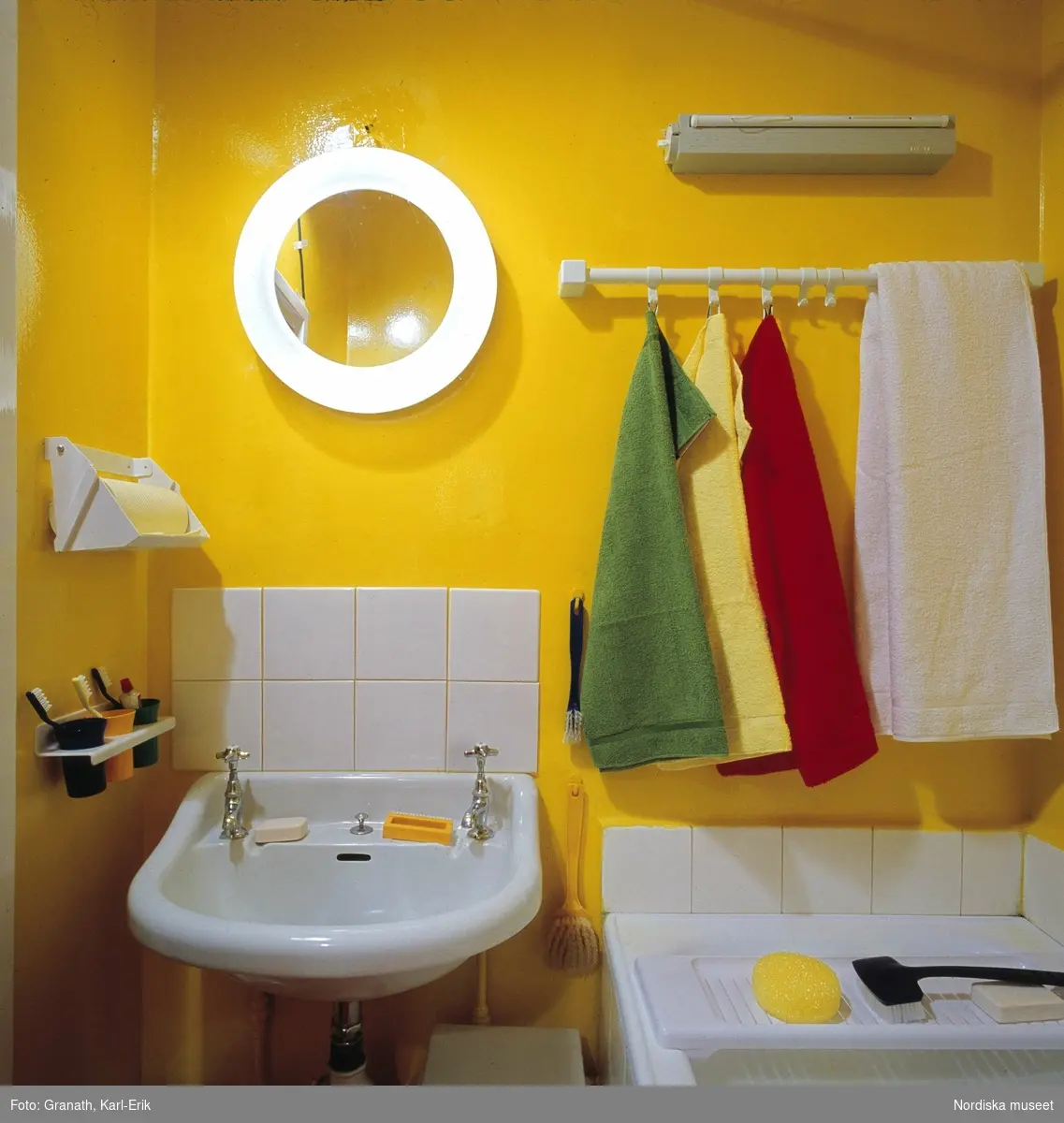Badrum med solgula väggar och vitt sanitetsporslin. Färgglada frottéhanddukar.
