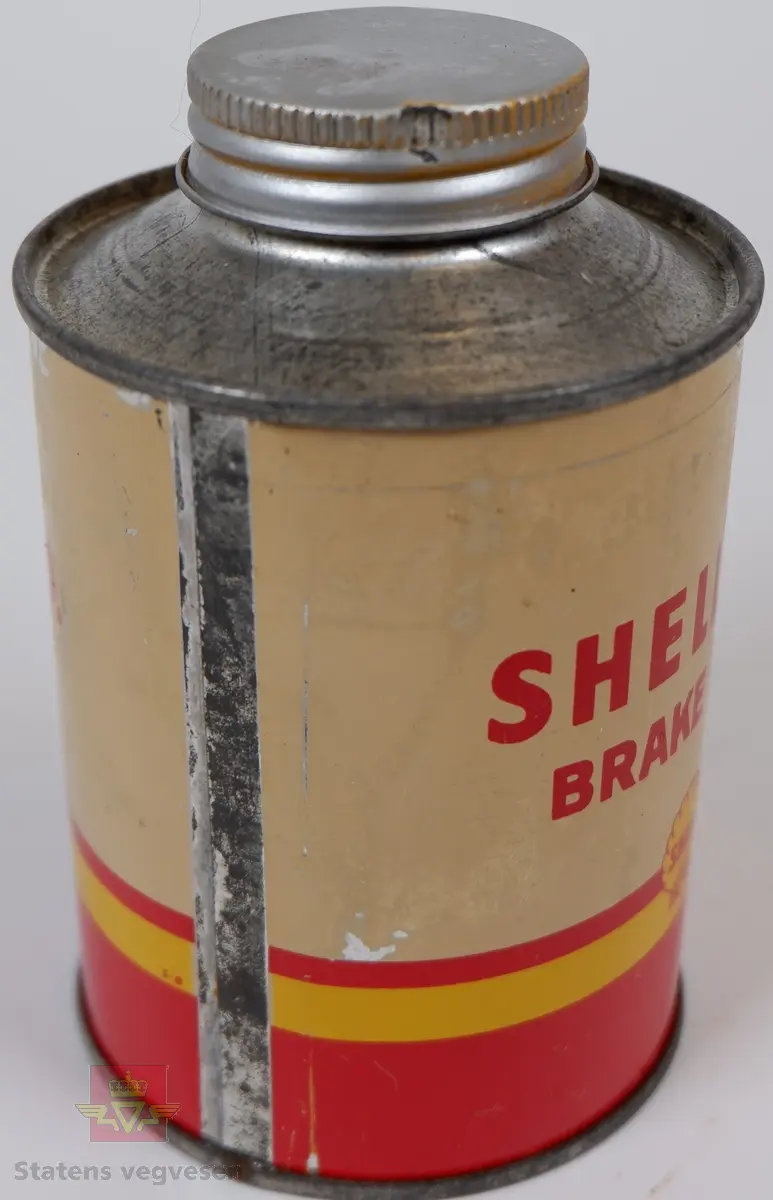 Rund utformet metallboks med skrukork. Den har en del rust, og lakk er stedvis flasset av. Boksen er tømt og rengjort.
Har inneholdt 0,5 liter bremsevæske. Lakkert hvit med rød nedre del. Påskrift med rødt: Shell H.D. brake fluid. Rødt felt har en gul stripe og Shell-logo i gult.
Ut i fra Shell-logoens utforming,  antas boksen å være fra cirka 1950-60. 
