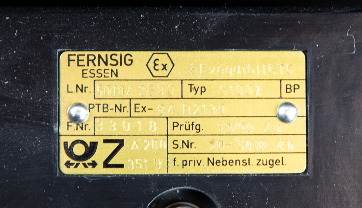 Telefon Fernsig Essen typ 5100. Rektangulär box i svart bakelit med cirkulär nummertavla. Telefonlur typ 2020 i svart bakelit med anslutningssladd i blått. Telefonen är brandskyddad och tillverkad så den inte avger någon gnistbildning.