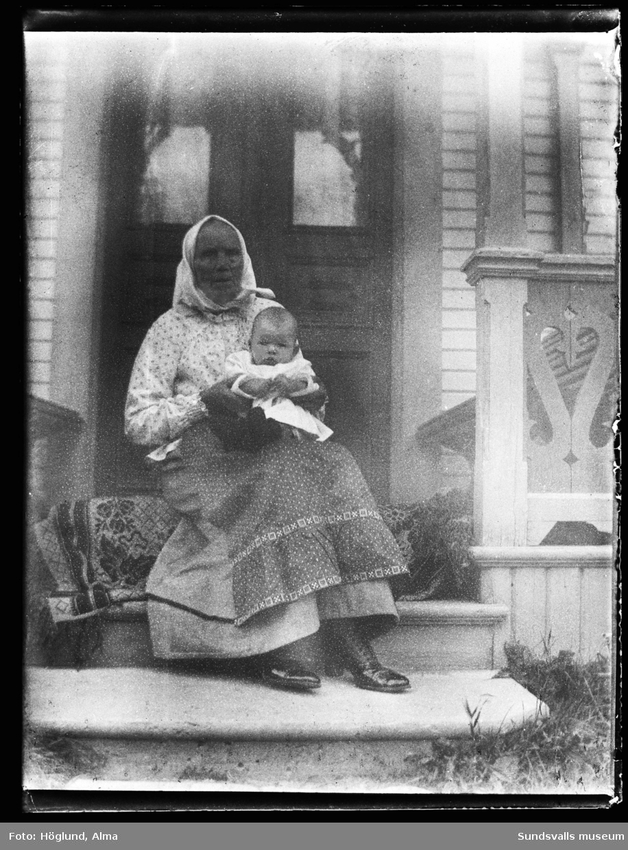 Porträttbild av en äldre kvinna med en baby i famnen sitter på brokvisten till Erik och Anna Wiströms hus i Fanbyn.