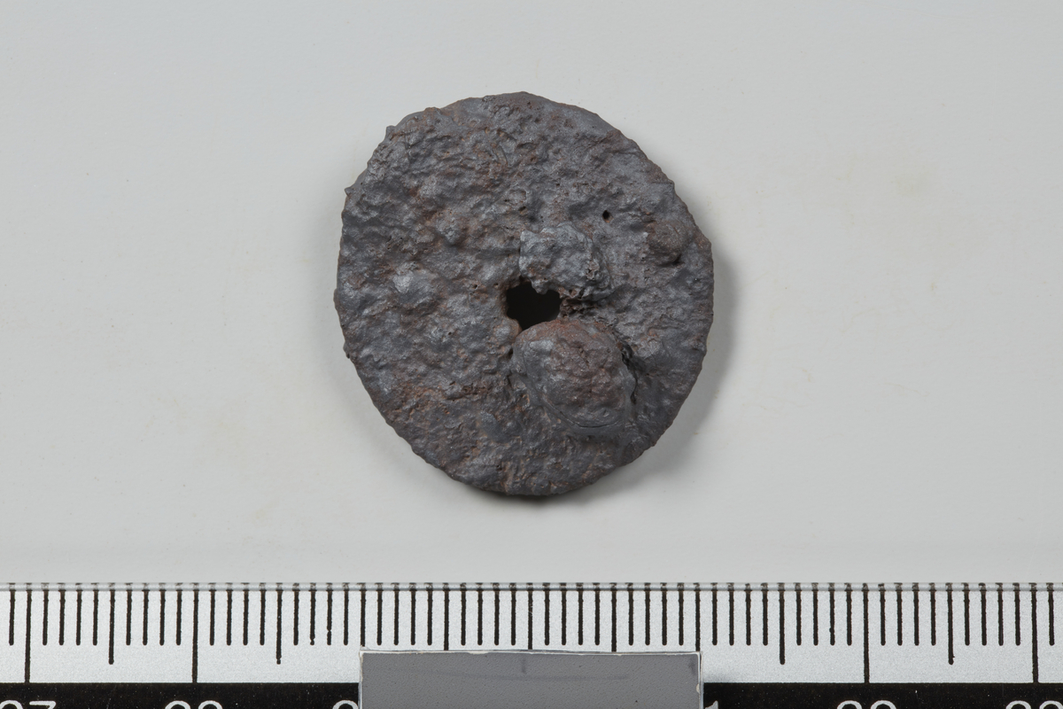 Rund skive av jern med hull i senter, mulig endeskive til kniv. L.2,2 cm, B.2,4 cm, T. 0,3 cm. Hullet har en diameter på 0,3 cm. Erodert.
