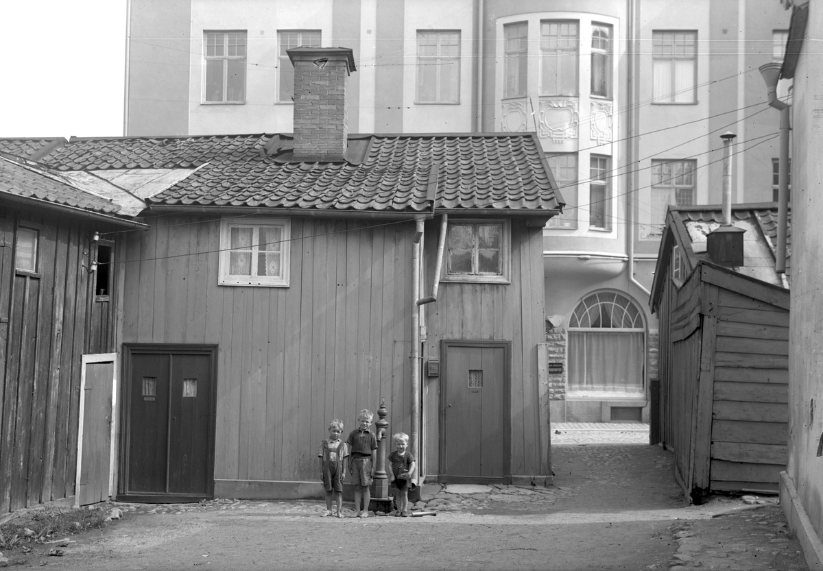 Innergården till Ågatan 15 i Linköping visar tydligt hur enkel bostadsstandarden var för många ännu under 1940-talet. Det verkar dock inte bekymrat de tre kamraterna invid pumpen särskilt mycket. Den höga fasaden i fonden tillhörde Weiders hus.