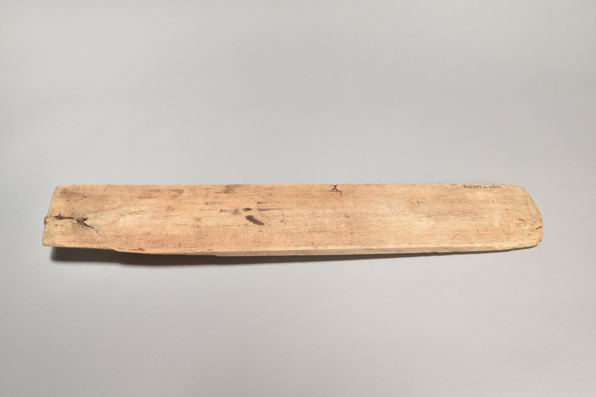 Kaveldon tillverkat i trä, med platt bräde och skuret handtag. Ovansidan av brädet med inskurna mönster. 