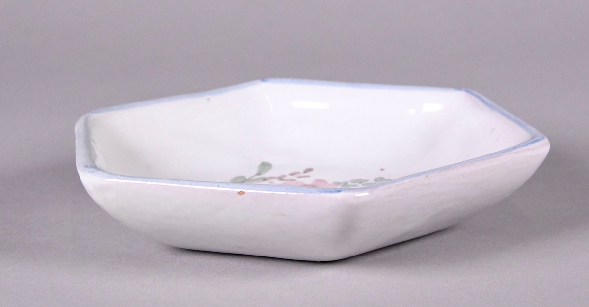 Skål av glassert keramikk, med opphøyd rand. Skålen er sekskantet, og har hvit grunnfarge. Håndmalt dekor av gule, røde og blå blomster, og en blå borde langs randen.