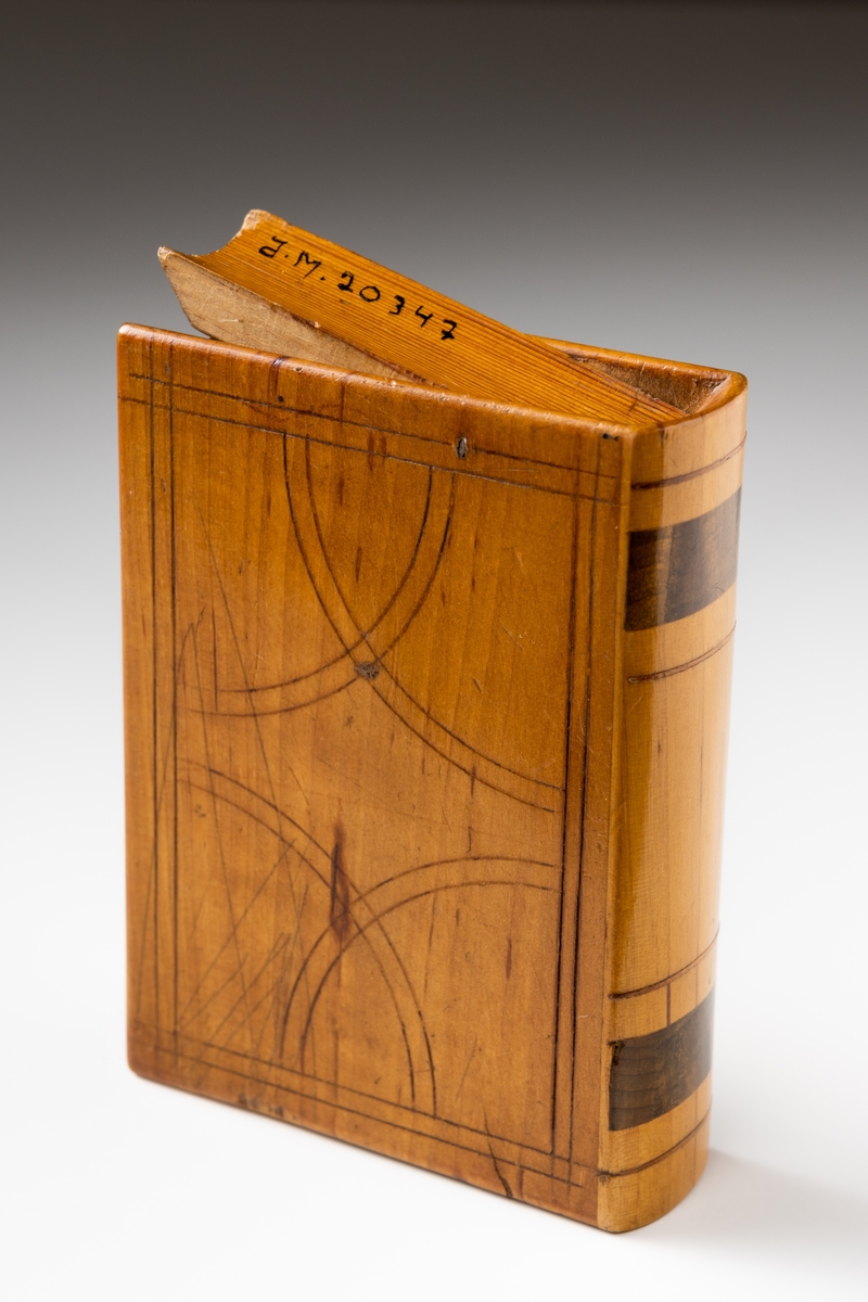 Sparbössa av trä i form av en bok dekorerad med ristat mönster och intarsia. På ovansidan av bössan finns en fällbar lucka innanför vilken myntinkastet är placerat. Skramlande föremål invändigt.