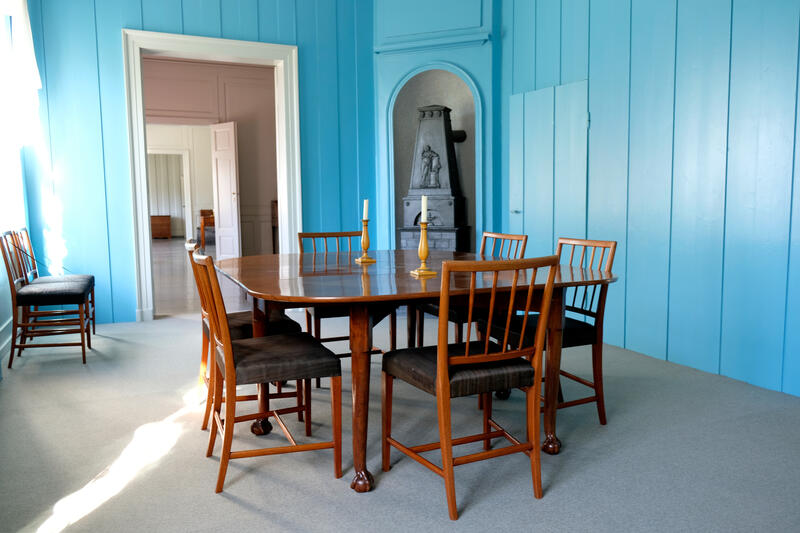 Konstitusjonskomiteens værelse i Eidsvollsbygningen har blåmalte plankevegger, grått gulv og i midten et stort mørkt trebord med stoler rundt. Veggene er nakne. I et hjørne står en ovn.