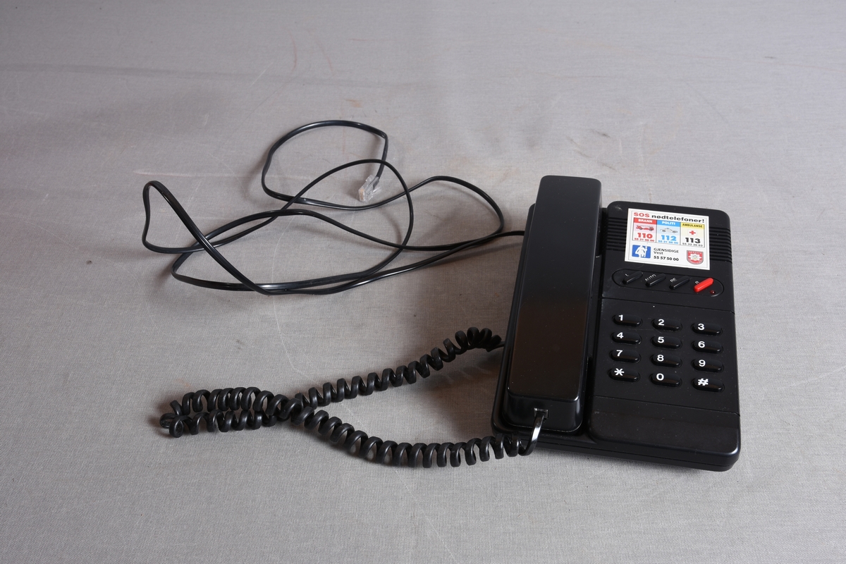 Svart fasttelefon med base, rør med ledning, samt tilkoblingskontakt.