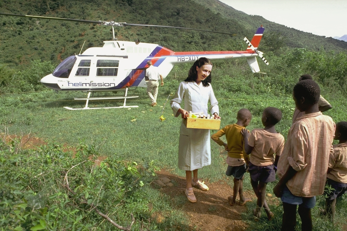 Kunstneren, kledd som hvitkledd sykepleier, har manipulert seg selv inn i et fotografi som viser et grønt landskap med afrikanske barn og et helikopter. 
Del av serie som består av digitale fotomontasjer hvor kunstneren plasserer seg selv i ulike roller som bistandsarbeider i Afrika og Asia.