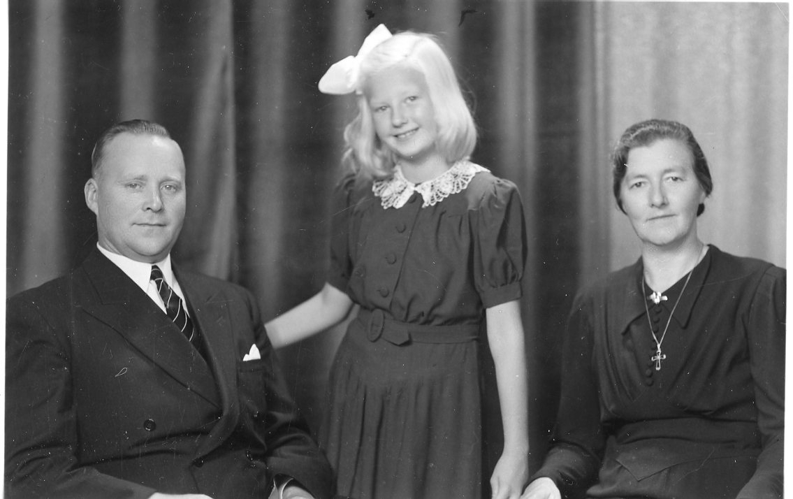 En ljushårig flicka med rosett i håret står mellan sin pappa och mamma.