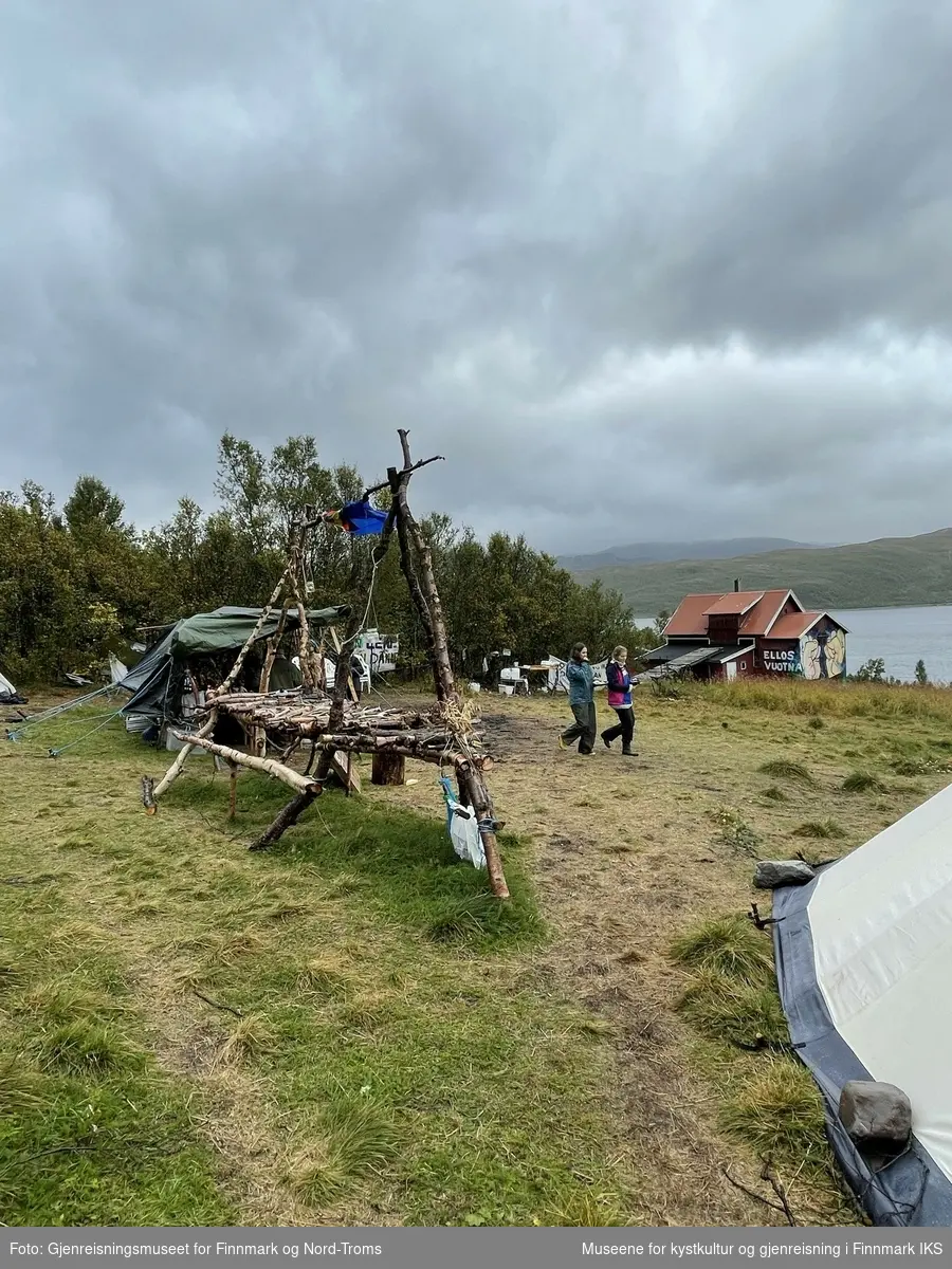 Protestleiren på Markoppneset i Finnmark i 2021. Protestbevegelsen mot dumping av gruveavfall i Repparfjorden har samlet seg og har etablert en teltleir. Bildet er del av en serie som dokumenterer leiren og omgivelsen i området. Alle avbildete personer har gitt sitt samtykke.
Bildet på naustveggen er et verk av kunstneren Tegson og har tittelen "Ellos vuotna".